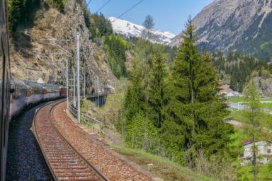 The Gotthard Panorama Express train south of Airolo in spring, with a beautiful combination of budding green nature and snowy mountain tops.