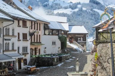 Medieval street in Gruyères against the backdrop of snowy forested hills.
